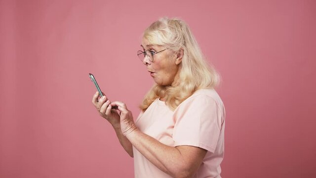 Modern senior woman browsing on cellphone and enjoying win, gesturing yes, pink studio background