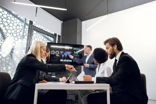 Senior professional businessman explains company's vision and financial infographics to his confident multiethnic diverse team, on digital wall board at joint meeting in boardroom.