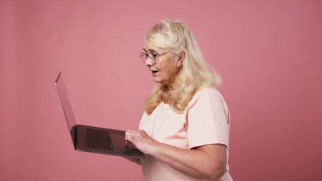 Happy excited senior woman in glasses working on laptop online and enjoying success, pink studio background