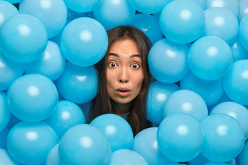Fototapeta na wymiar Close up shot of brunette Asian woman stares at camera stucks head through blue inflated balloons poses over decorated background expresses amazement going to celebrate birthday. Holiday event