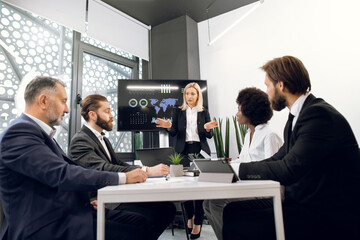 Attractive confident blond mature lady in formal suit, gesturing and reporting about ecological, economic or marketing problems to her multiethnic team of colleagues in boardroom during meeting