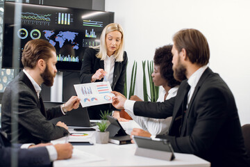 Close up of attractive confident experienced blond female leader, showing infographics and financial charts on the paper during meeting to her focused professional multiracial partners