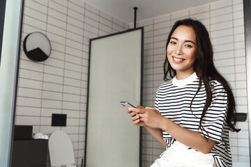 Fototapeta na wymiar Young asian woman sitting on bathtub with smartphone, smiling at camera. Pretty girl checking mobile app in bathroom