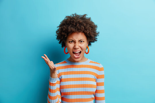 Beautiful dark skinned woman with curly hair raises hand and shouts loudly stands mad irritated expresses anger rage wears striped casual jumper isolated over blue background. Negative emotions
