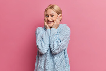 Positive blonde blue eyed young European woman keeps hands on cheeks and smiles pleasantly...