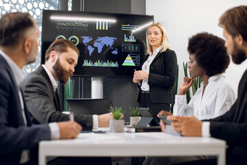 Middle aged pleasant confident blond business lady, showing company's work infographics on digital wall screen during meeting with team of diverse multiracial male and female businesspeople
