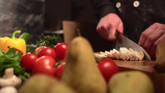 A man chef in a black jacket cuts cheese on a cutting board among fresh vegetables. Close-up of hands. Slider shot.