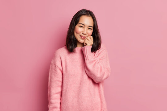 Lovely brunette woman with eastern appearance keeps hand on face has gentle smile looks satisfied at camera has dimples on rouge cheeks poses against pink background. Cute glad Asian girl indoor