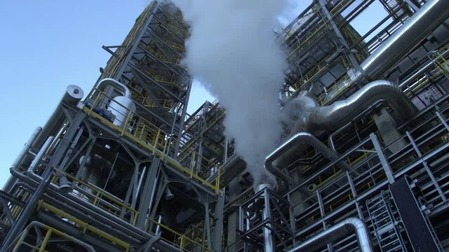A Low Angle Shot Of Smoke From An Oil Refinery. The challenges of reducing greenhouse gas emissions and air pollution through energy sources.  Interlacing of metal pipes