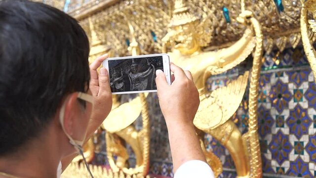 New normal lifestyle Attractive senior Asian traveler wearing mask protective prevent COVID-19 using smartphone taking photo at Wat Pra Kaew Temple of Buddha in Bangkok Thailand, Tourist joyful camera