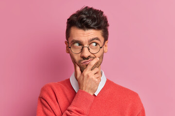 Handsome thoughtful man holds chin and looks doubtful away purses lips makes planning tries to...