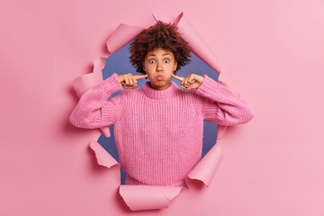 Funny dark skinned woman makes grimace holds breath and points index fingers on blowing cheeks keeps eyes widely opened wears knitted sweater breaks through pink paper background. Face expressions