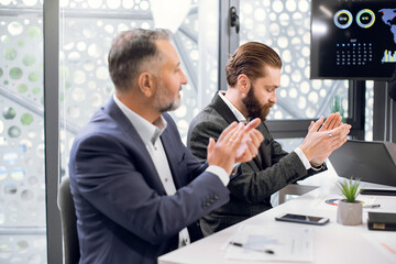 Good speech, business, congratulation and appreciation concept. Two satisfied European diverse business men in formal wear, sitting at the table in conference hall and applauding. Focus on young man