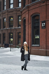 a young woman stands in a light warm outerwear in winter near a brick loft building with tall black windows