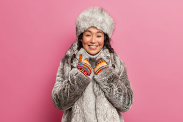 Pretty young woman with eastern appearance dressed in grey fur coat smiles sincerely hears something pleasant going to have winter walk poses against pink background. Eskimo from arctic place