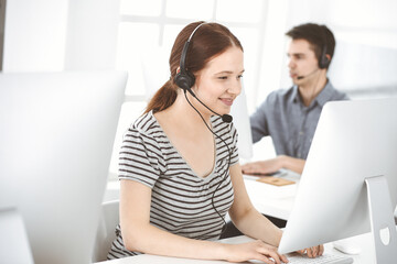 Casual dressed young woman using headset and computer while talking with customers online. Group of operators at work. Call center, business concept