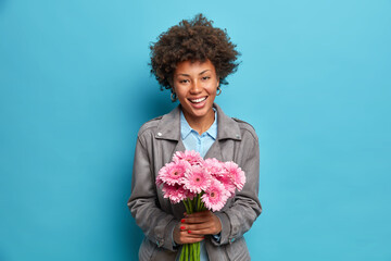 Studio shot of happy culy haired woman holds beautiful flowers receives her favorite gerberas from...