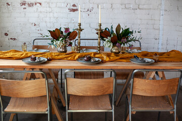 long wooden party table with stylish decoration, with candlesticks, vases of flowers, plates and food in a room with a white brick wall