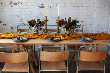long wooden party table with stylish decoration, with candlesticks, vases of flowers, plates and food in a room with a white brick wall