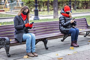 Two young women wearing medical masks talking on a cell phone at a safe social distance while sitting on a bench in an autumn park. Meeting and chatting with friends during the coronavirus pandemic