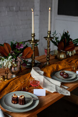 long wooden party table with stylish decoration, with candlesticks, vases of flowers, glasses with wine, name cards, plates and food in a room with a white brick wall