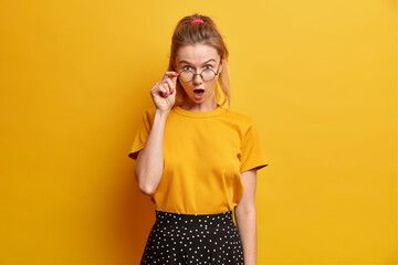 Indoor shot of astonished European girl stares with widely opened mouth keeps hand on rim of...