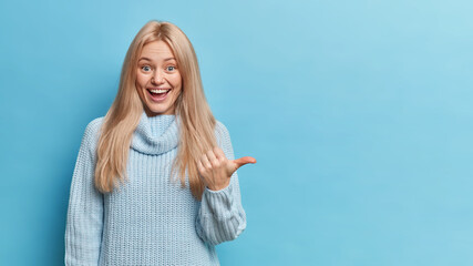 Beautiful glad blonde woman points thumb on right wears knitted sweater shows blank space for your promotional content poses against blue background. Monochrome shot. Place advert banner there