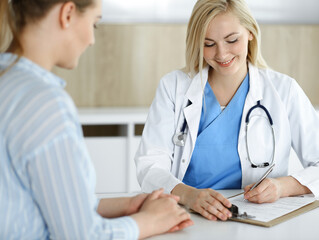 Fototapeta premium Woman-doctor and patient sitting and talking in hospital or clinic. Blonde therapist is cheerfully smiling. Medicine concept