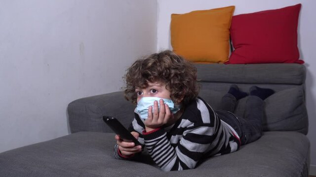 Europe, Italy , MIlan - Child boy 6 years old watching television and playing video games with mask during Covid-19 Coronavirus lockdown quarantine home 