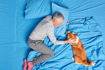 Photo of happy aged bearded man in nightwear plays with dog expresses love and care pose together on comfortable bed have friendly relationships Male pensioner enjoys lazy day with pet at home