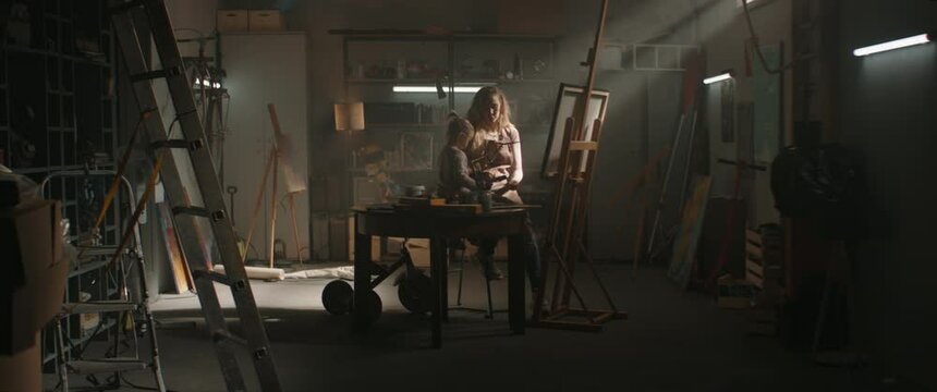 WIDE Female painter artist working in studio inside her garage, little daughter helping with the picture. Shot with 2x anamorphic lens
