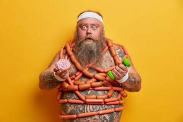 Stunned bearded overweight adult man has imbalanced nutrition has big tattoed belly wrapped with sausages tries to go in for sport poses surprised against yellow background. Junk food concept