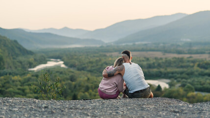 Young man hugs his girlfriend on the mountain peak at sunset background.