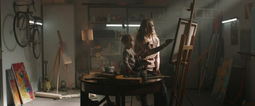 WIDE Female painter artist working in studio inside her garage, little daughter helping with the picture. Shot with 2x anamorphic lens