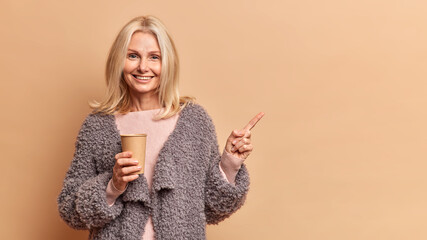 Studio shot of lovely blonde fifty years old woman smiles positively holds disposable paper cup of...