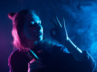 The girl smokes VAPE. A woman with a VAPE makes an OK sign with her hand. Vaper girl on a dark...