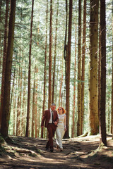 Happy stylish couple newlyweds in the green forest on summer day. bride in long white dress and groom in red suit are hugging. wedding day.