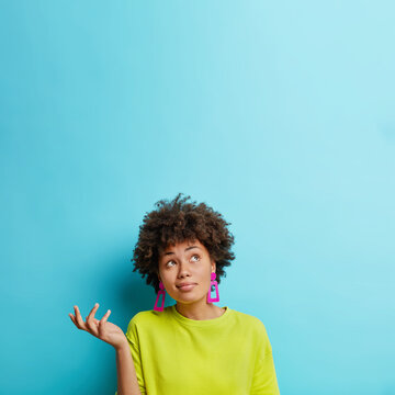 Vertical shot of doubtful Afro American woman raises hand with doubt looks confused above makes decision dressed casually isolated over blue background blank space for your promotional content