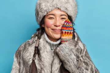 Cheerful tender Eskimo woman wears winter clothes keeps hand on cheek enjoys winter time smiles gladfully poses against blue background. Cold climate. Inuit female adapted to frosty weather. - obrazy, fototapety, plakaty