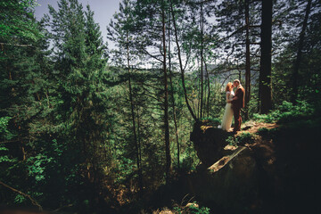 Happy stylish couple newlyweds in the green forest on summer day. bride in long white dress and groom in red suit are hugging. wedding day.