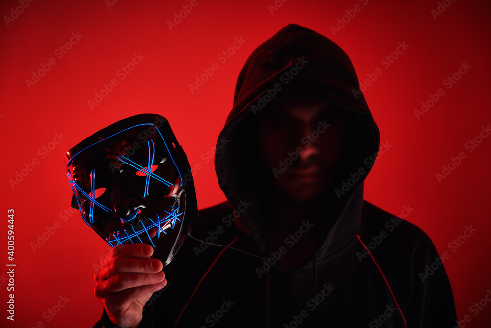 Wall mural Man in hoodie with blurred face hold neon glowing scary mask. Anonymous concept