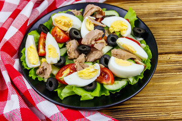 Tasty tuna salad with lettuce, black olives, eggs and fresh vegetables on wooden table