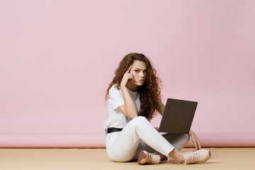 Beautiful curly-haired girl in casual white clothes sits on the floor.  Working from home. Pastel trendy pink background. Sad woman holding laptop and looking at camera.