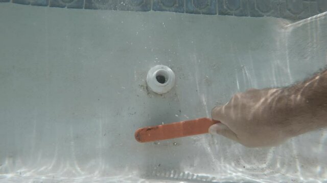 A underwater video of a brush scrubbing dirt and algae from the side of a pool and water outlet