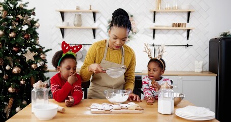 Happy African American family with little cute kids on Christmas standing at table in kitchen and decorate homemade baked xmas gingerbread cookies New Year preparations, winter season holidays concept