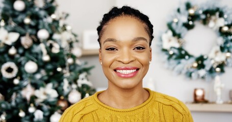 Cheerful handsome African American female looking at camera and smiling indoors near Christmas glowing decorated tree in good mood feeling happy.