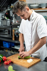 Young male chef in uniform bending over table while cutting fresh green zucchini