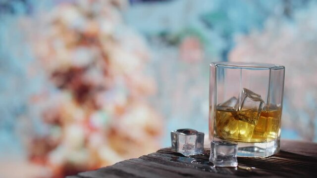 A glass of whiskey with ice is on a wooden table. In the background there is a New Year's landscape, there is a New Year tree and snow is slowly falling.