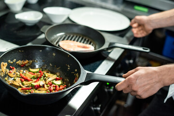 Hands of male chef roasting piece of salmon and vegetable stew on frying pans