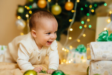 Cute smiling baby is lying under a festive Christmas tree and playing with gifts. Christmas and New Year celebrations